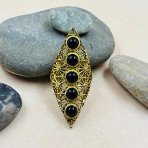 Puede incluir: Un anillo alargado de color dorado con un diseño geométrico. El anillo presenta cinco piedras negras redondas engastadas en un marco en forma de diamante. El marco tiene un patrón de filigrana.