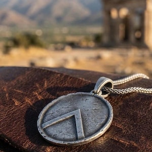 May include: A circular silver-toned metal pendant featuring a raised inverted V-shaped emblem on a textured, weathered surface. The pendant is attached to a silver-toned woven chain, showcasing a distressed finish with visible surface markings.