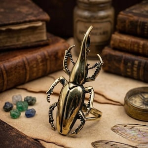 May include: A metallic gold-toned ring featuring a large, detailed beetle design with a prominent horn and articulated legs. The beetle is crafted with a polished finish, showcasing segmented body parts and a band underneath for wearing on a finger.