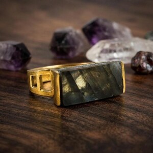 Può includere: Un anello color oro con una pietra rettangolare in labradorite grigio scuro con visibili riflessi iridescenti. La fascia presenta un design geometrico traforato con intagli quadrati sul lato, una finitura metallica e una superficie piatta e lucida.