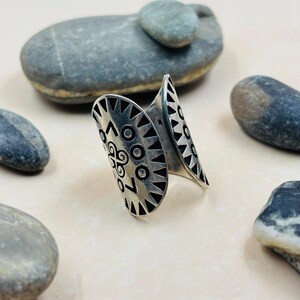 Puede incluir: Un brazalete plateado con un diseño abierto. El brazalete presenta patrones geométricos, incluyendo triángulos y círculos. El brazalete se exhibe sobre una superficie clara con piedras grises. El diseño del brazalete incluye líneas curvas.