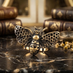 Op de afbeelding: Een goudkleurige metalen ring met een groot, gedetailleerd bijenornament. De bij heeft een gepolijste kop en borststuk, terwijl de vleugels en het achterlijf zijn versierd met een herhalend patroon van cirkelvormige uitsparingen.