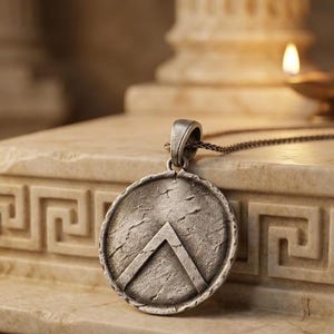 May include: A circular silver-toned pendant featuring a raised inverted V-shaped emblem on a textured, weathered surface. The pendant is attached to a thin metal chain, showcasing a metallic finish with dark, recessed details across the face.