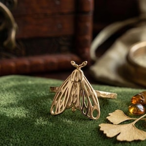 May include: A gold-toned ring featuring a detailed moth design with open-work wings and a textured band. The moth is centered on the ring, showcasing symmetrical wing patterns and small antennae. The metal has a polished finish with visible light reflections.