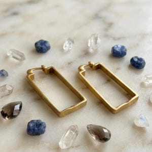 May include: A pair of gold-toned rectangular hoop earrings with a matte finish, featuring a hinged closure mechanism at the top. The earrings are displayed alongside several loose, small, multi-colored gemstones including clear, blue, and dark brown pieces.
