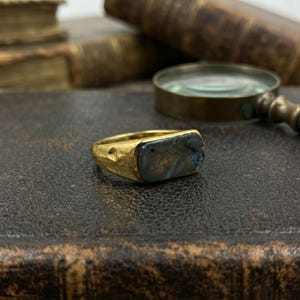 May include: A gold ring featuring a rectangular, dark, iridescent stone set into a textured, hammered metal band. The stone displays subtle flashes of blue and gold colour, while the band has a rustic, uneven finish with a visible indentation.