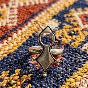 May include: A silver-toned ring featuring a prominent, vertically oriented centerpiece. The design consists of a circular top section connected to a diamond-shaped base, with small protruding points at the sides and a tiny sphere at the bottom.