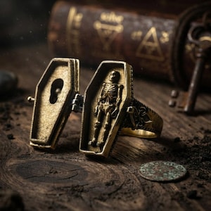 May include: A gold-toned metal ring featuring a hinged coffin design that opens to reveal a sculpted skeleton figure inside. The exterior of the coffin lid displays a keyhole cutout, and the band of the ring has intricate side detailing.