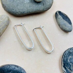 May include: A pair of silver-colored, elongated oval hoop earrings. The earrings are displayed on a light beige surface, with several gray stones scattered around them. The earrings have a simple, minimalist design.