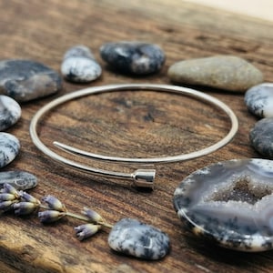 May include: A silver-toned bypass bangle bracelet featuring a thin, circular wire band that overlaps at the front. The ends of the bracelet are finished with small, cylindrical caps. The design is simple, sleek, and metallic in appearance.
