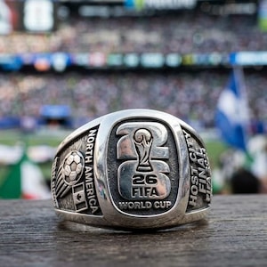 Könnte beinhalten: Ein silberner Ring mit dem Text "26 FIFA WORLD CUP" und einem Fußball-Design. Der Ring trägt auch die Aufschrift "NORTH AMERICA" und "HOST CITY FINAL". Im Hintergrund ist ein Stadion mit einer Menschenmenge zu sehen.