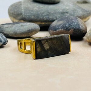 May include: A gold-colored ring with a rectangular, dark gray stone. The ring's band has a geometric design with cut-out squares. The stone has a textured appearance, and the ring is set against a neutral background with gray stones.