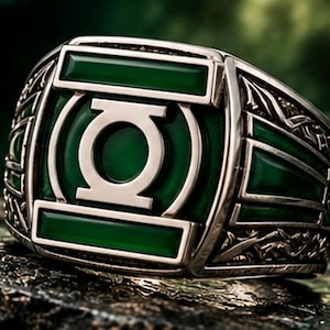 May include: A silver-toned ring featuring a prominent circular Green Lantern emblem centered on a rectangular green inlay. The band is detailed with intricate metalwork patterns on the sides, framing the central green stone and metallic logo.