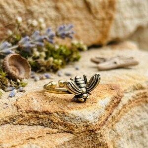 Op de afbeelding: Een goudkleurige metalen ring met een gedetailleerd bijenontwerp als middelpunt. De bij heeft gestructureerde vleugels en een gesegmenteerd lichaam, bevestigd aan een eenvoudige, dunne, open ring. De ring is gemaakt van metaal met een gepolijste afwerking.