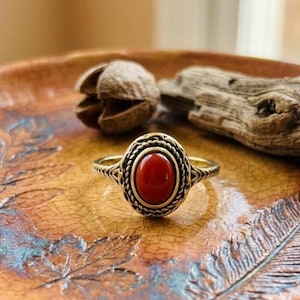 Puede incluir: Un anillo de tono dorado con una piedra roja central de forma ovalada. El engaste está decorado con un borde de metal trenzado similar a una cuerda y patrones de filigrana intrincados a lo largo de la banda, creando un aspecto detallado.