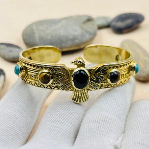 May include: Gold-toned cuff bracelet featuring an eagle design with turquoise, tiger's eye, and black onyx stones. The open-ended bracelet has an engraved pattern and is approximately 2.5 inches in diameter.