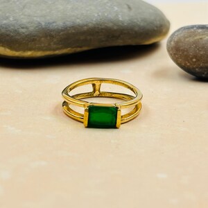 Op de afbeelding: Een goudkleurige ring met een rechthoekige, smaragdgroene edelsteen. De ring heeft een dubbele band, met de edelsteen in het midden. De ring wordt gepresenteerd op een lichtgekleurd oppervlak.