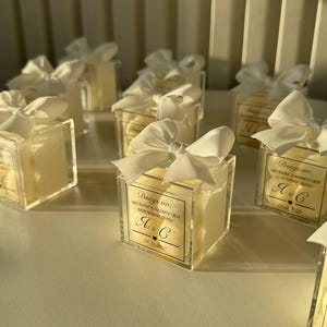 Puede incluir: Varias cajas de acrílico transparente, cada una con un artículo de color claro y atada con un lazo de cinta blanco. Cada caja tiene una etiqueta con texto y las iniciales "A & C" y una fecha. Las cajas están dispuestas sobre una superficie clara.