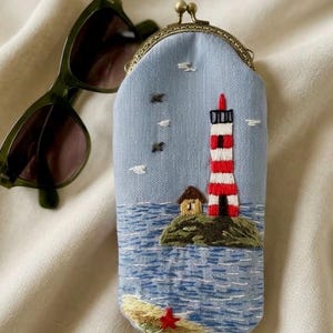 May include: A light blue fabric pouch with a metal kiss-lock clasp, featuring an embroidered scene of a red and white striped lighthouse on a rocky island, a small house, blue ocean waves, a sandy shore with a red starfish, and dark sunglasses.