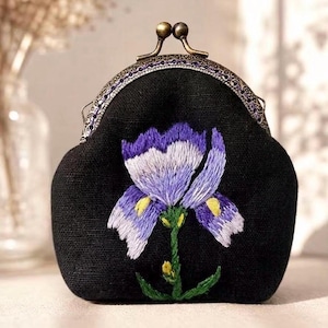 May include: A black coin purse featuring an embroidered purple and white iris flower with green stems. The purse has a textured fabric exterior and a metallic bronze-toned kiss-lock clasp with an intricate engraved pattern along the frame edge.