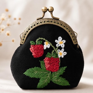 May include: A black coin purse with a bronze-toned metal kiss-lock clasp. The front features hand-embroidered red raspberries, green leaves, and small white flowers with yellow centers. The fabric has a visible weave texture.