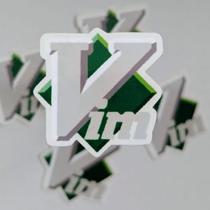 May include: A die-cut sticker featuring the text Vim in a bold, light grey, sans-serif font with a subtle drop shadow. The letters are layered over a dark green diamond shape with a lighter green border, creating a geometric, layered appearance.