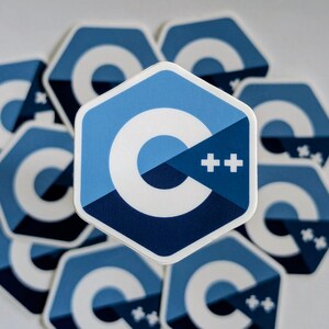 May include: A hexagonal sticker featuring the C++ programming language logo. The design displays a large white letter C on a blue background, with two white plus signs positioned to the right. The sticker has a white border and a geometric shape.