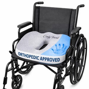 May include: A gray and blue orthopedic seat cushion with a handprint design, placed on a black wheelchair. The cushion has a central cutout and the text "ORTHOPEDIC APPROVED" and "Comfy Friend".