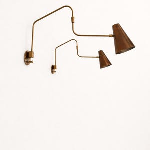 Puede incluir: Dos apliques de pared ajustables con una est&eacute;tica vintage. Las l&aacute;mparas presentan un acabado de lat&oacute;n con pantallas c&oacute;nicas en un tono cobre c&aacute;lido. El dise&ntilde;o incluye brazos articulados para iluminaci&oacute;n direccional.