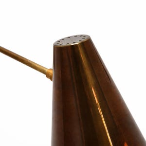 Puede incluir: Primer plano de un cabezal de l&aacute;mpara de forma c&oacute;nica, de estilo vintage. El cabezal de la l&aacute;mpara es de color cobre c&aacute;lido y bru&ntilde;ido, con un brazo de lat&oacute;n que se extiende hacia la izquierda. La parte superior del cabezal de la l&aacute;mpara tiene varios agujeros peque&ntilde;os. El fondo es blanco.