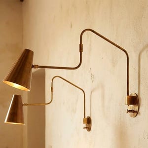 Puede incluir: Dos apliques de pared de lat&oacute;n con pantallas c&oacute;nicas y brazos ajustables est&aacute;n montados en una pared texturizada. Los apliques tienen una est&eacute;tica vintage, con un brillo c&aacute;lido y acogedor que emana de las pantallas.