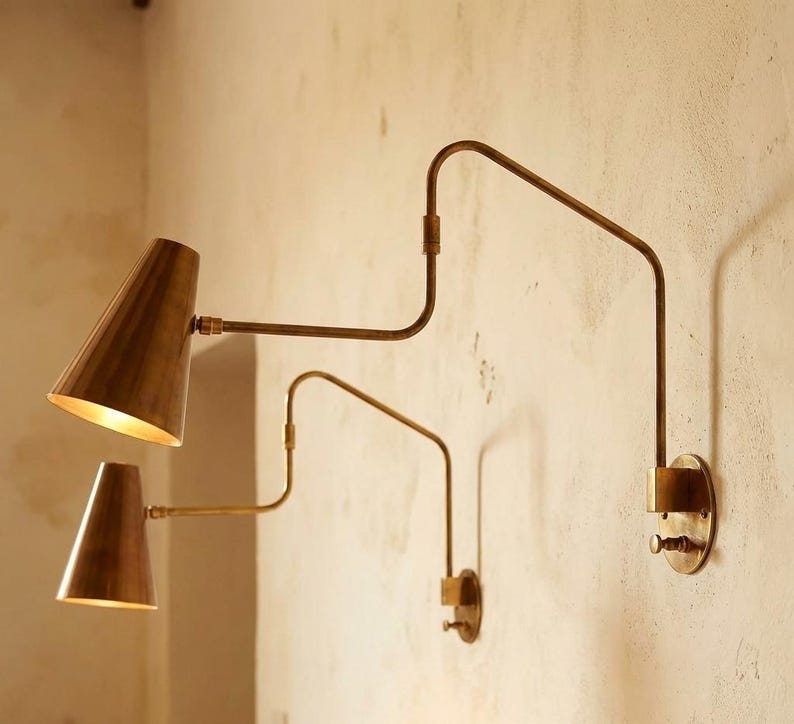 Puede incluir: Dos apliques de pared de lat&oacute;n con pantallas c&oacute;nicas y brazos ajustables est&aacute;n montados en una pared texturizada. Los apliques tienen una est&eacute;tica vintage, con un brillo c&aacute;lido y acogedor que emana de las pantallas.