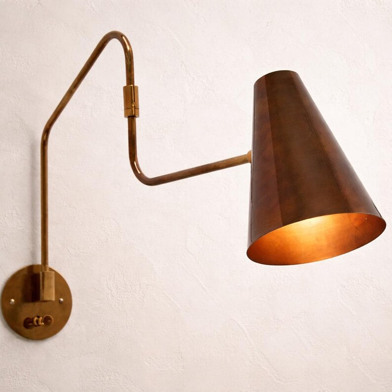 Puede incluir: Aplique de pared en tono lat&oacute;n con una pantalla c&oacute;nica de color cobre oscuro. La fuente de luz es visible en el interior. La l&aacute;mpara tiene un brazo ajustable y una base redonda con un interruptor.
