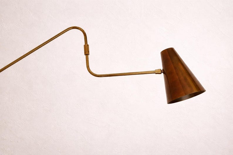 Puede incluir: Una l&aacute;mpara de pared de estilo vintage, con acabado en lat&oacute;n. Presenta un brazo largo y curvado y una pantalla c&oacute;nica en un tono cobre c&aacute;lido. El dise&ntilde;o es minimalista y elegante, adecuado para varios estilos de interior.