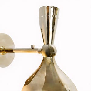 May include: A brass wall sconce with a cone-shaped shade and a cylindrical arm. The sconce has a round base and a small, decorative detail on the arm. The finish is a polished gold color.