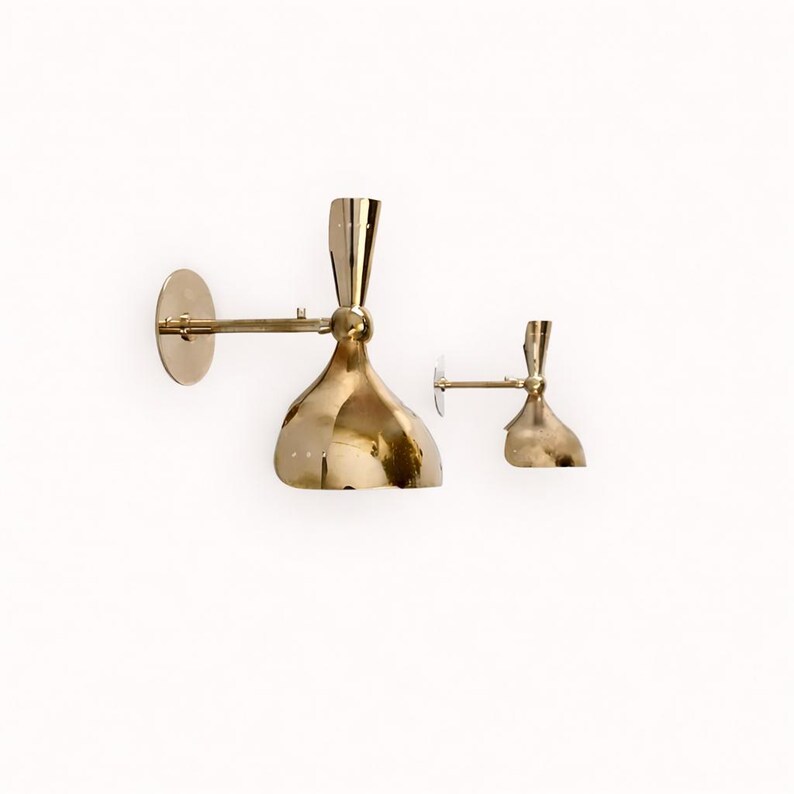 May include: Two brass wall sconces with a unique, curved design. The sconces have a gold finish and are mounted on a white wall. The larger sconce has a circular backplate and an adjustable arm. The smaller sconce is a miniature version.