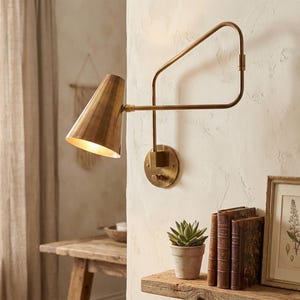 Puede incluir: Una l&aacute;mpara de pared de lat&oacute;n con una pantalla c&oacute;nica y un brazo ajustable. Est&aacute; montada en una pared texturizada. Una peque&ntilde;a planta suculenta en maceta y una obra de arte enmarcada se encuentran en una estanter&iacute;a de madera debajo.