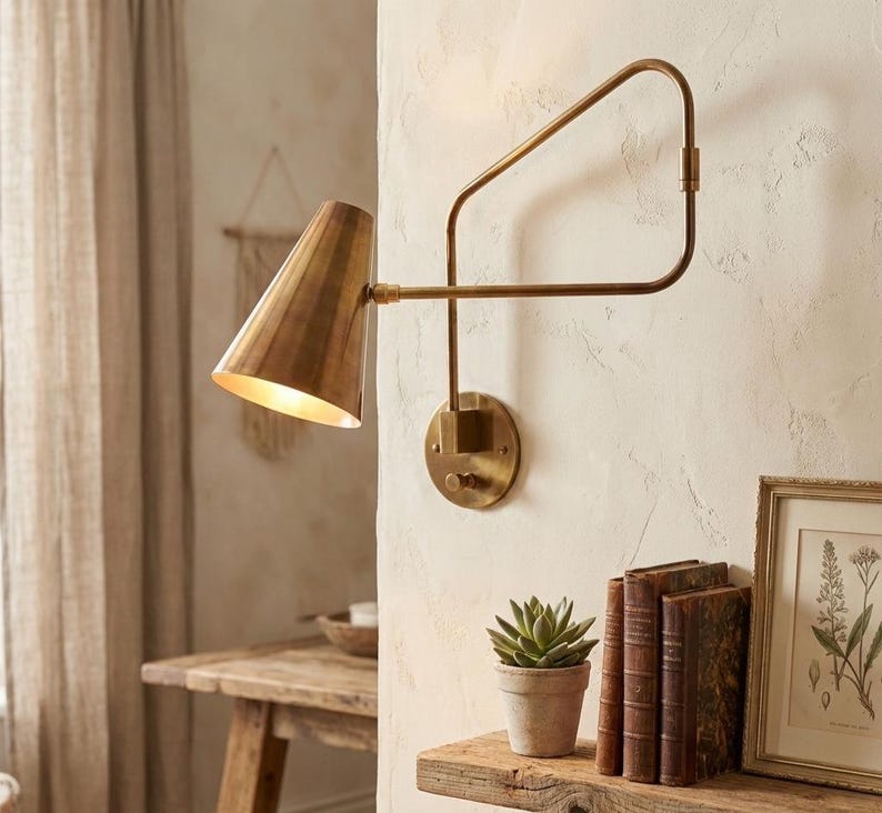 Puede incluir: Una l&aacute;mpara de pared de lat&oacute;n con una pantalla c&oacute;nica y un brazo ajustable. Est&aacute; montada en una pared texturizada. Una peque&ntilde;a planta suculenta en maceta y una obra de arte enmarcada se encuentran en una estanter&iacute;a de madera debajo.