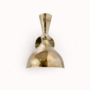 May include: A brass-colored wall sconce with a conical top and a rounded, bell-shaped shade. The sconce is mounted on a circular base and features a series of small, decorative rivets. The design is vintage and elegant.
