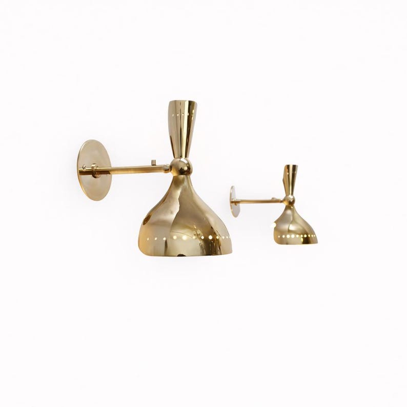 May include: Two gold-colored wall sconces with a mid-century modern design. Each sconce features a round wall plate, a horizontal arm, and a bell-shaped shade with small, evenly spaced holes. The sconces are made of a shiny, reflective metal.