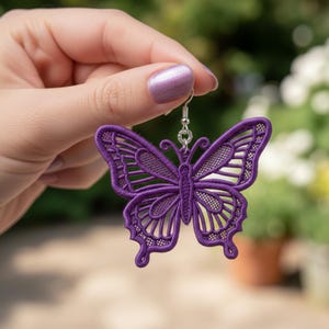 Pode incluir: Um brinco de borboleta roxa com detalhes bordados intrincados. O brinco apresenta um gancho prateado e um design delicado e vazado. As asas da borboleta são de um tom vibrante de roxo, mostrando as costuras detalhadas.