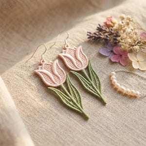 May include: Embroidered tulip earrings in soft pink and green. The earrings feature a detailed floral design with a delicate, textured appearance. The earrings are displayed on a neutral fabric background, alongside a pearl necklace and dried flowers.