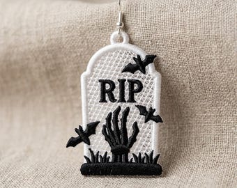 Puede incluir: Un pendiente colgante con forma de lápida bordada sobre un fondo de malla blanca con detalles en hilo negro. El diseño incluye el texto RIP, una mano esquelética emergiendo del suelo y tres pequeños murciélagos negros volando.