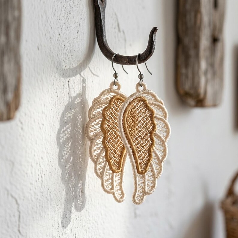 May include: A pair of dangling earrings shaped like angel wings. The earrings are made of beige and gold-colored lace, with a detailed, openwork design. They hang from silver-colored hooks, creating a delicate and ethereal aesthetic. The earrings are displayed against a white wall.