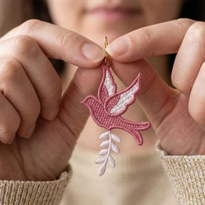May include: A pink embroidered dove earring with white wing details and a sprig of leaves. The dove is suspended from a gold-colored hook. The earring is held up by a person.