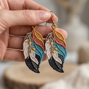May include: A pair of dangle earrings featuring embroidered feather shapes in shades of mustard yellow, terracotta, teal, cream, and black. The intricate threadwork creates a textured leaf-like pattern with small circular accents, suspended from bronze-toned metal hooks.