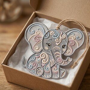 K&ouml;nnte beinhalten: Graues Elefanten-Ornament mit aufwendigen Stickereidetails in Hellblau, Rosa und Creme. Der Elefant liegt in einer braunen Schachtel mit wei&szlig;em Papierfutter und einer nat&uuml;rlichen Schnurschlaufe zum Aufh&auml;ngen.