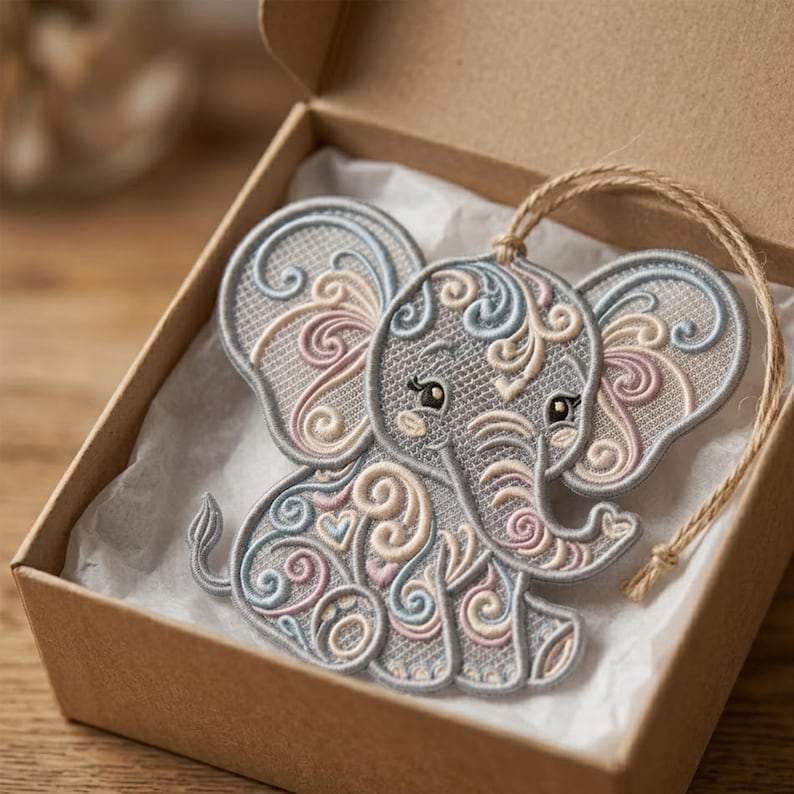 K&ouml;nnte beinhalten: Graues Elefanten-Ornament mit aufwendigen Stickereidetails in Hellblau, Rosa und Creme. Der Elefant liegt in einer braunen Schachtel mit wei&szlig;em Papierfutter und einer nat&uuml;rlichen Schnurschlaufe zum Aufh&auml;ngen.