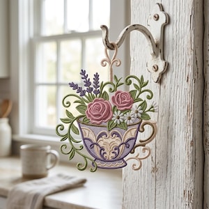 Può includere: Decorazione da parete a forma di tazza da tè ricamata con rose rosa, lavanda e margherite. La tazza da tè è viola e crema con dettagli ornati. È appesa a un gancio di metallo bianco.