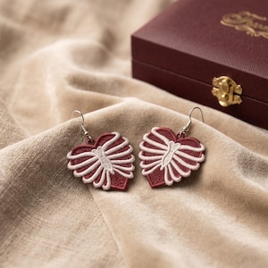 May include: A pair of heart-shaped earrings featuring embroidered red fabric with a white stitched ribcage design. Each earring is attached to a silver-toned metal hook for hanging, showcasing detailed needlework in a contrasting color scheme.